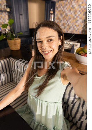Smiling woman taking selfie in kitchen, sitting on striped bench Smiling woman taking selfie in kitchen, sitting on striped bench 116316050