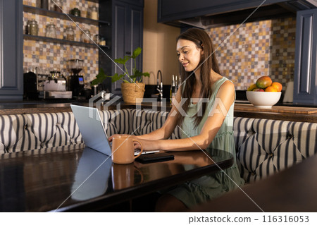Working from home, young woman using laptop at kitchen table 116316053