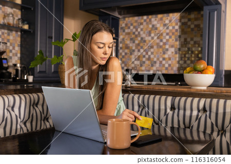 Using credit card and laptop, woman shopping online in cozy kitchen setting 116316054