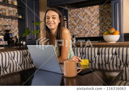 Using laptop and holding credit card, woman smiling in kitchen with coffee mug Using laptop and holding credit card, woman smiling in kitchen with coffee mug 116316063