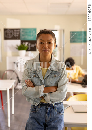 Teenage girl in high school classroom standing with arms crossed, looking confident 116316076