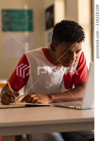 In high school, teenage boy writing in notebook while using laptop in classroom 116316109