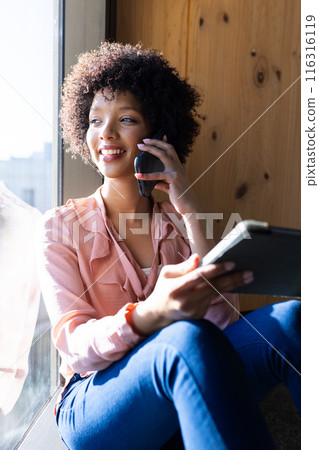 Talking on smartphone and holding tablet, woman smiling by window in office 116316119
