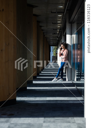 Standing in corridor, woman drinking coffee and using smartphone, enjoying break, copy space 116316123