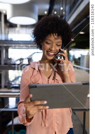 Smiling woman using tablet and talking on smartphone in modern office Smiling woman using tablet and talking on smartphone in modern office 116316140