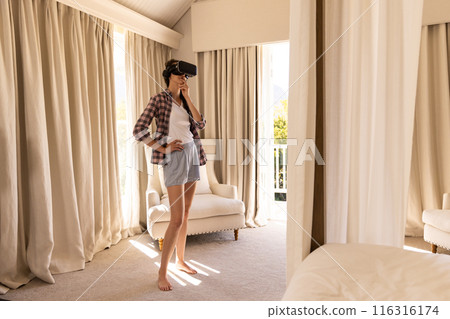 Using VR headset, young woman exploring virtual reality in living room 116316174