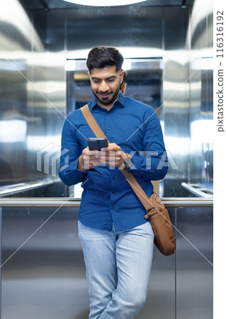 Using smartphone, man in blue shirt standing in elevator with brown bag 116316192
