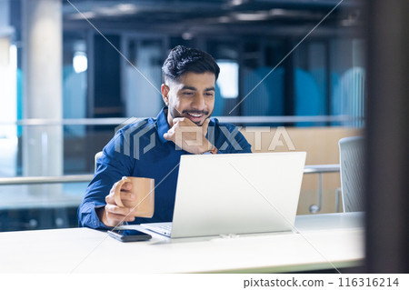 Smiling businessman holding coffee cup and using laptop at office desk 116316214