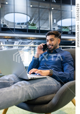 Working from office, man using laptop and talking on smartphone, smiling, copy space Working from office, man using laptop and talking on smartphone, smiling, copy space 116316239