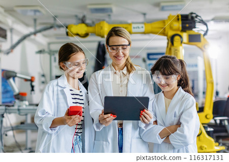 Female teacher with two schoolgirls in lab coats, standing in front robotic arm. After-school robotics club. Children learning robotics in Elementary school. Girls in science. Female teacher with two schoolgirls in lab coats, standing in front robotic arm. After-school robotics club. Children learning robotics in Elementary school. Girls in science. 116317131