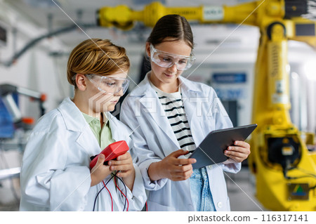 Two classmates standing in robotic laboratory, wearing lab coats and safety eyeglasses. After-school robotics club. Children learning robotics in Elementary school. 116317141