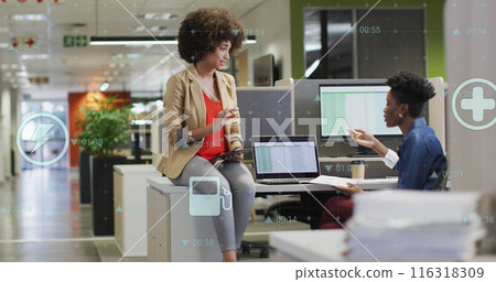 Image of multiple digital icons against two diverse women discussing at office 116318309