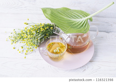 Herbal medicinal tea with chamomile flowers in a glass teapot, a bouquet of fresh flowers, on a wooden light background. 116321398