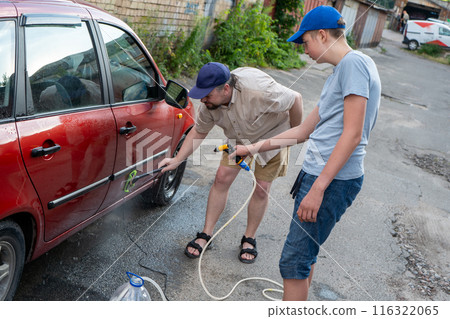 Dad and teenage son work together on the street near the garage. Outdoor activity. Family spend time. Helping and teach. Washing the car with a hand pump and hose. Bottled water. Dad and teenage son work together on the street near the garage. Outdoor activity. Family spend time. Helping and teach. Washing the car with a hand pump and hose. Bottled water. 116322065