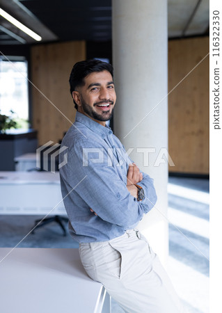 Smiling businessman in office, leaning on desk with arms crossed Smiling businessman in office, leaning on desk with arms crossed 116322330