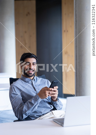 Smiling businessman using smartphone while sitting at desk with laptop in office Smiling businessman using smartphone while sitting at desk with laptop in office 116322341
