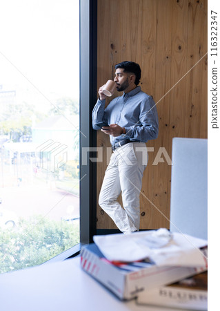 Drinking coffee and using smartphone, businessman standing by office window 116322347