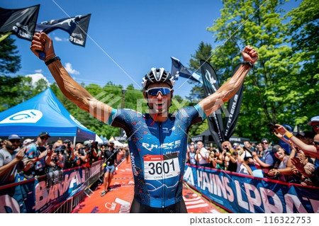 A cyclist raises his arms in victory as he crosses the finish line of a race, surrounded by cheering spectators. 116322753