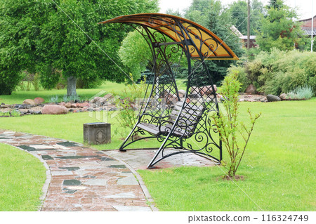 Wrought iron swing with canopy in park near stone path. Recreation in nature. Seasons and enjoyment.  116324749