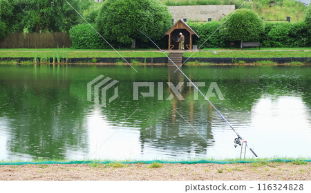 Fishing lake with wooden arbour for relaxing. Fishing rods and equipment in summer. Fishing lake with wooden arbour for relaxing. Fishing rods and equipment in summer. 116324828