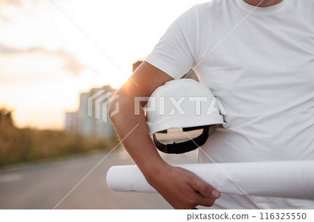 Unrecognisable male civil engineer holds protective white hard hat and blueprint on multi-apartment residential complex backdrop. Real estate concept. Industry new building business. 116325550
