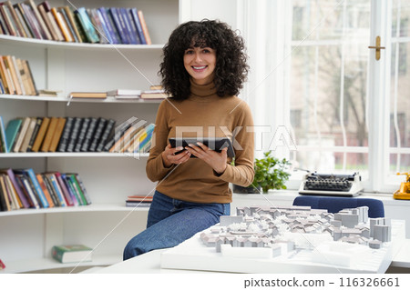 Brunette woman with a tablet in hands looking contented while working in a library Brunette woman with a tablet in hands looking contented while working in a library 116326661