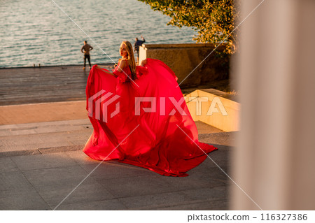 Sunrise red dress. A woman in a long red dress against the backdrop of sunrise, bright golden light of the sun's rays. The concept of femininity, harmony. 116327386