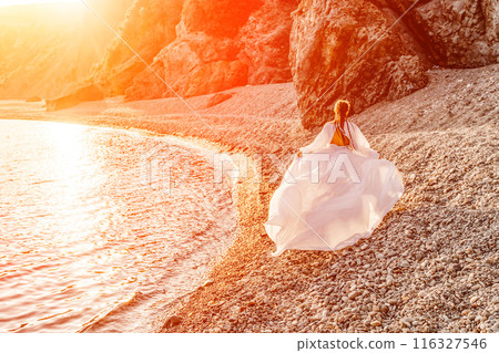 Mysterious woman silhouette long hair walks on the beach ocean water, sea nymph wind listens to the wave. Throws up a long white dress, a divine sunset. Artistic photo from the back without a face 116327546