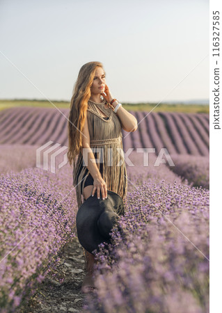 A woman is standing in a field of purple flowers, wearing a black dress and a black hat. 116327585