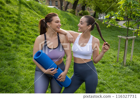 Active women friends wearing sporty attires holding mat preparing for park yoga workout together Active women friends wearing sporty attires holding mat preparing for park yoga workout together 116327731