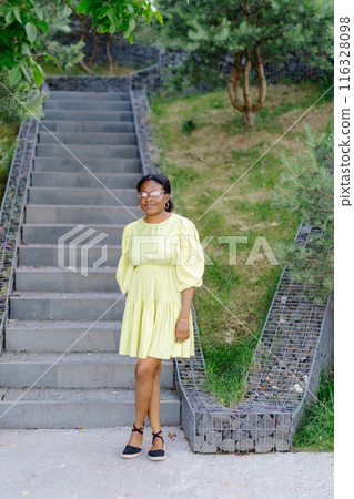 Dark-skinned lady laughs outside. Brunette woman in yellow dress smiles and walks outdoors Dark-skinned lady laughs outside. Brunette woman in yellow dress smiles and walks outdoors 116328098