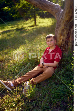 preteen boy in a red t-shirt is sad near a withered tree in the park 116328669