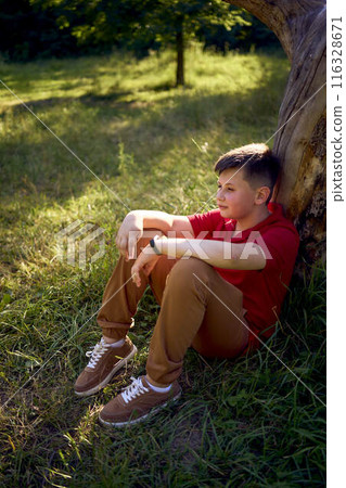 preteen boy in a red t-shirt is sad near a withered tree in the park 116328671