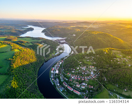 A sunset aerial shot of Orlik Reservoir in Czechia shows a winding river, hills, and a quaint town in the valley with vibrant blue and golden skies. 116334314