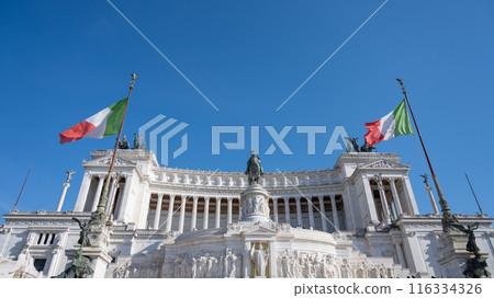 The Victor Emmanuel II National Monument in Rome, Italy, is adorned with Italian flags waving against a blue sky. The massive, white marble structure stands tall, a testament to the country history. The Victor Emmanuel II National Monument in Rome, Italy, is adorned with Italian flags waving against a blue sky. The massive, white marble structure stands tall, a testament to the country history. 116334326