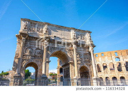 The Arch of Constantine, a Roman triumphal arch dedicated to Emperor Constantine, stands against a clear blue sky. The archs ornate carvings and intricate details are visible. 116334328