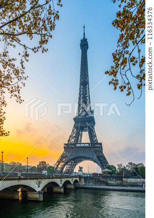 A view of the Eiffel Tower at sunrise in Paris, France. The iconic structure stands tall against a vibrant sky, with a bridge in the foreground and a river flowing through the city. 116334376