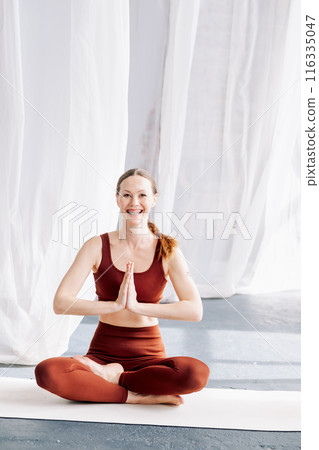 A slender woman with braces in a brown sports uniform sits in the lotus position against a background of white curtains and meditates. Yoga classes in the loft studio A slender woman with braces in a brown sports uniform sits in the lotus position against a background of white curtains and meditates. Yoga classes in the loft studio 116335047
