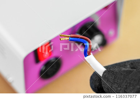 Electric cable with bare exposed wires and copper terminals in the hand of an electrician in front of the connecting power machine with control panel 116337077
