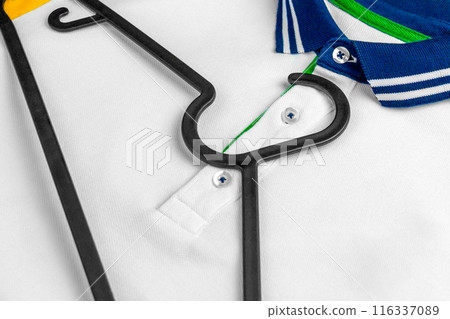White polo shirt with a blue green collar and yellow sleeves and a black plastic hanger lying on top. Stylish casual sportswear. Copy space White polo shirt with a blue green collar and yellow sleeves and a black plastic hanger lying on top. Stylish casual sportswear. Copy space 116337089