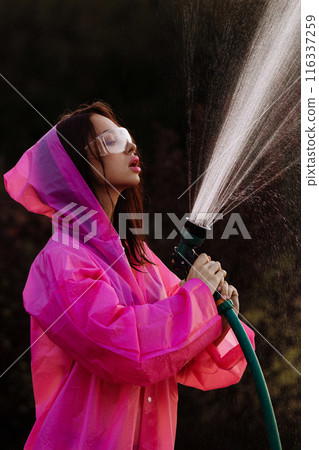 A glamour woman in a pink raincoat is spraying water from a hose A glamour woman in a pink raincoat is spraying water from a hose 116337259