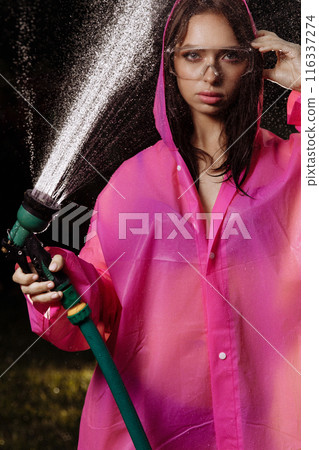 A glamour woman in a pink raincoat is spraying water from a hose 116337274