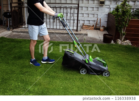 A young man is mowing a lawn with a lawn mower in his beautiful green floral summer garden 116338938