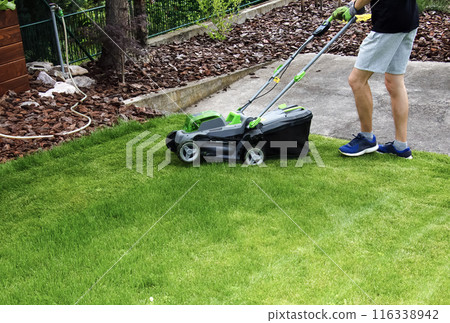 A young man is mowing a lawn with a lawn mower in his beautiful green floral summer garden A young man is mowing a lawn with a lawn mower in his beautiful green floral summer garden 116338942