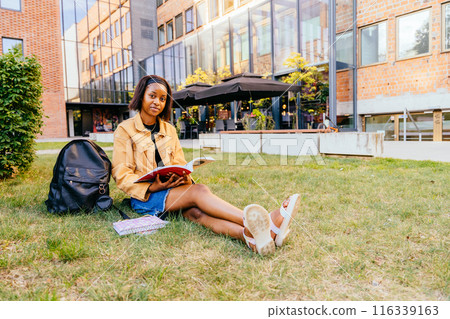 African woman sits on the grass with a book in her lap. She is wearing a yellow jacket and blue jeans. The scene is set in front of a building with a large window African woman sits on the grass with a book in her lap. She is wearing a yellow jacket and blue jeans. The scene is set in front of a building with a large window 116339163