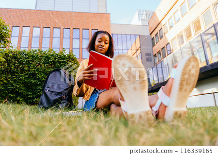 A young student black ethnic girl sitting on grass with a book in her hand. She is smiling and enjoying her time. The book is open to a page, and there is a backpack nearby. Scene suggests a relaxed A young student black ethnic girl sitting on grass with a book in her hand. She is smiling and enjoying her time. The book is open to a page, and there is a backpack nearby. Scene suggests a relaxed 116339165