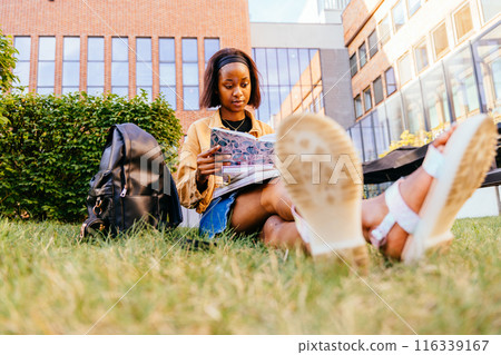 Adorable young African American woman reading a good book while chilling on grass lawn in university park. 116339167