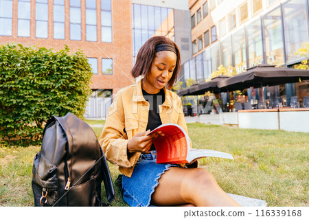 Young African American female student reading books cheerfully while sitting on grass area university campus with building in the background. 116339168