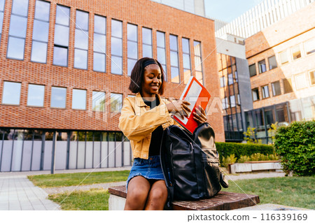 A young woman is sitting on a bench with backpack and exercise book. She is smiling and she is enjoying her time. Concept of relaxation and leisure A young woman is sitting on a bench with backpack and exercise book. She is smiling and she is enjoying her time. Concept of relaxation and leisure 116339169