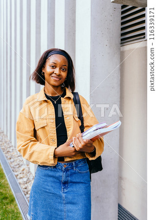 A young woman wearing a yellow jacket and blue jeans is holding a stack of books. She has a smile on her face and she is happy A young woman wearing a yellow jacket and blue jeans is holding a stack of books. She has a smile on her face and she is happy 116339171